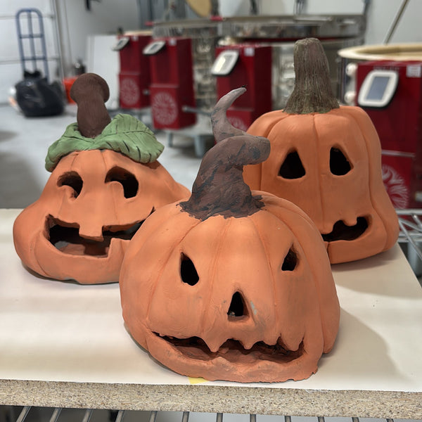 Three handbuilt clay jack-o'-lantern pumpkin lanterns with carved faces on a studio shelf at Claya Studios, RiNo, Denver