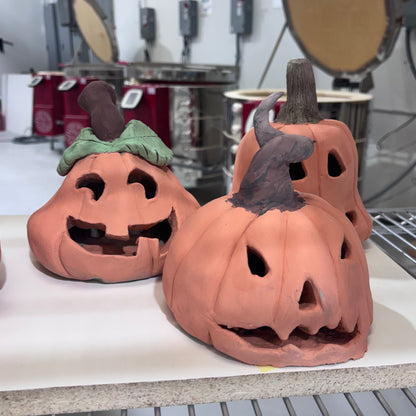 Handbuilt clay pumpkin lanterns, one with a curled witch hat stem and one with a green leaf, in the kiln room at Claya, Denver