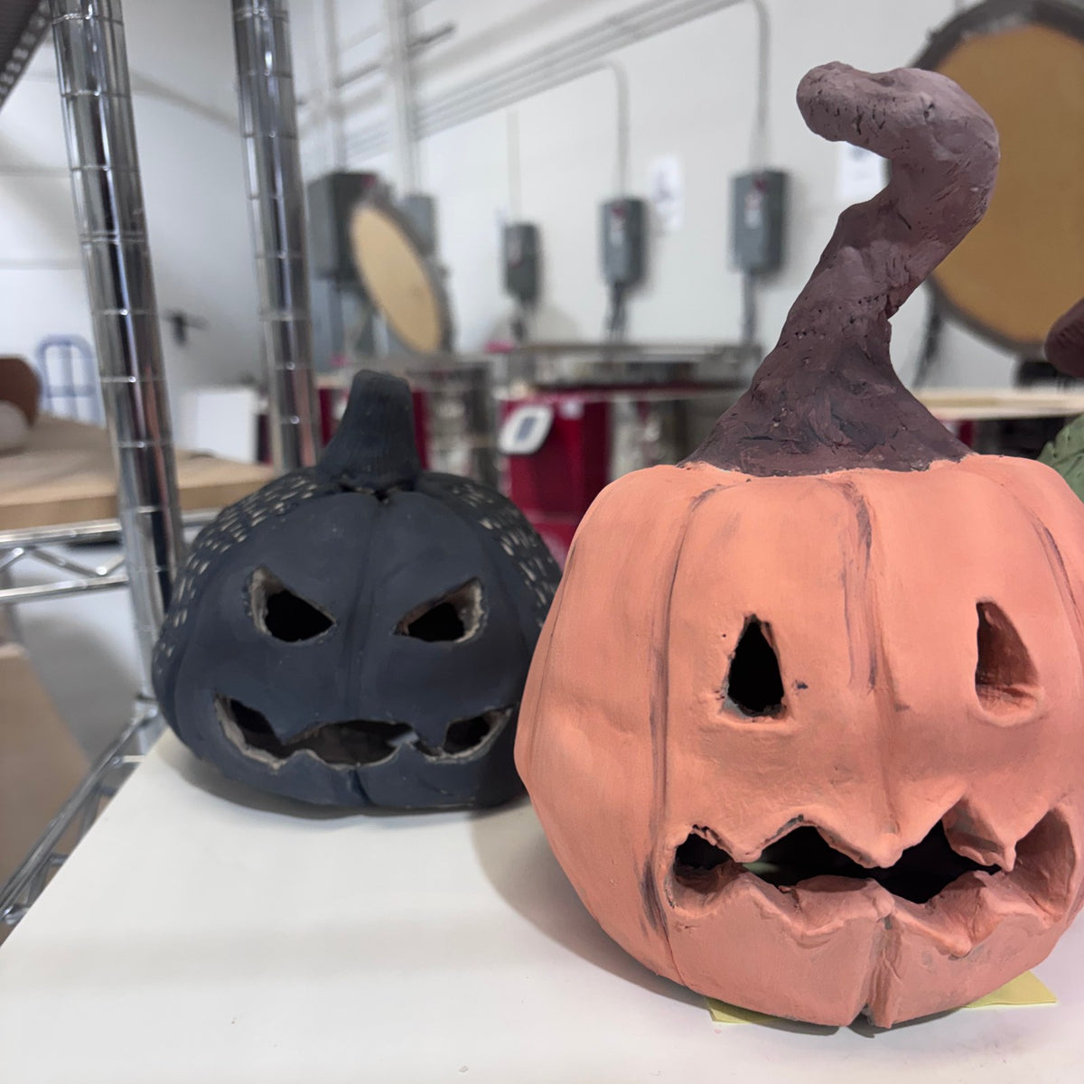 A black clay pumpkin lantern next to a terracotta jack-o'-lantern with a curly stem, handbuilt at Claya Studios in Denver