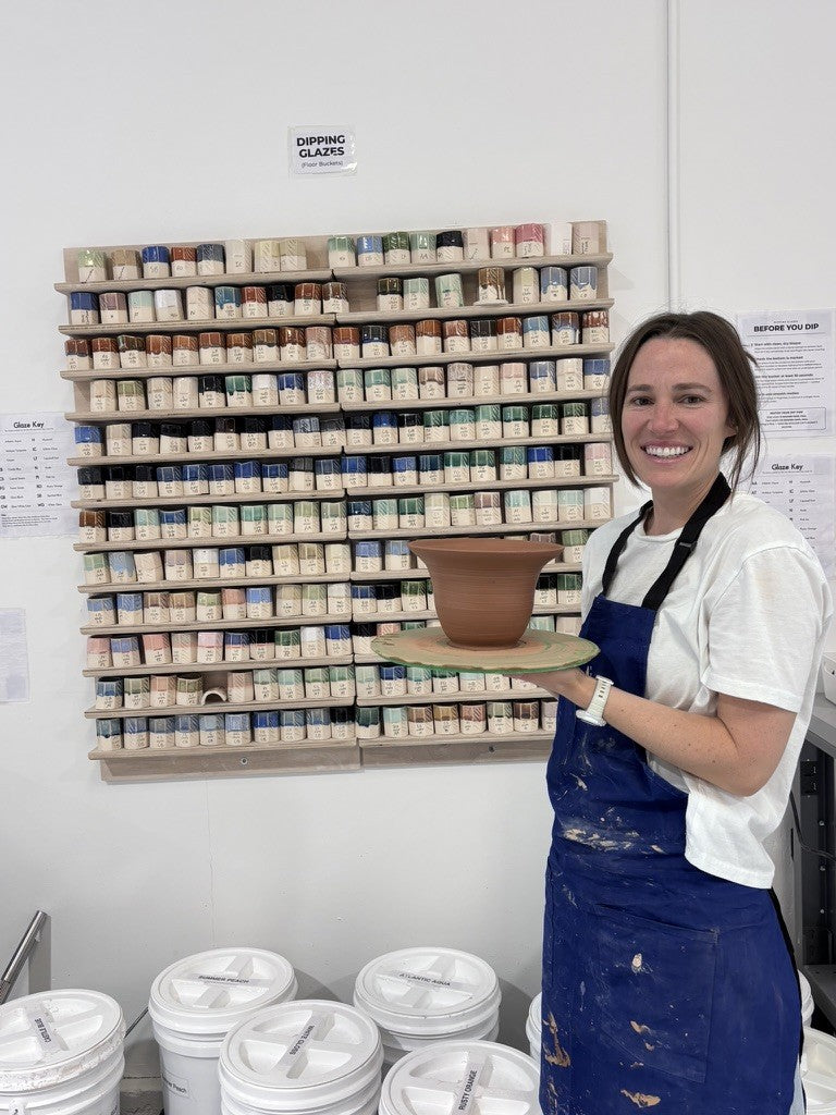 Smiling student in a blue apron holding a thrown clay bowl in front of the glaze wall at Claya Studios in RiNo, Denver