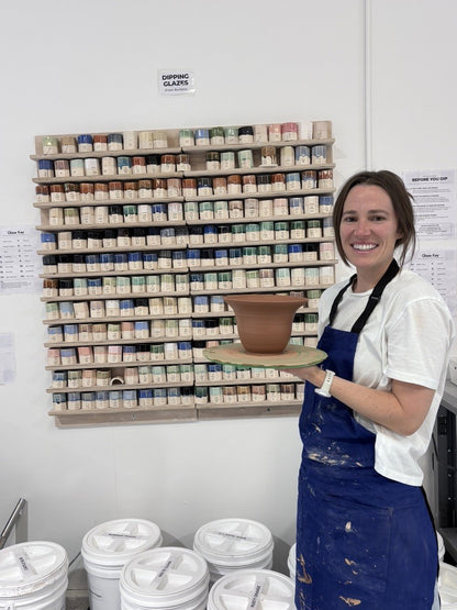 Smiling student in a blue apron holding a thrown clay bowl in front of the glaze wall at Claya Studios in RiNo, Denver